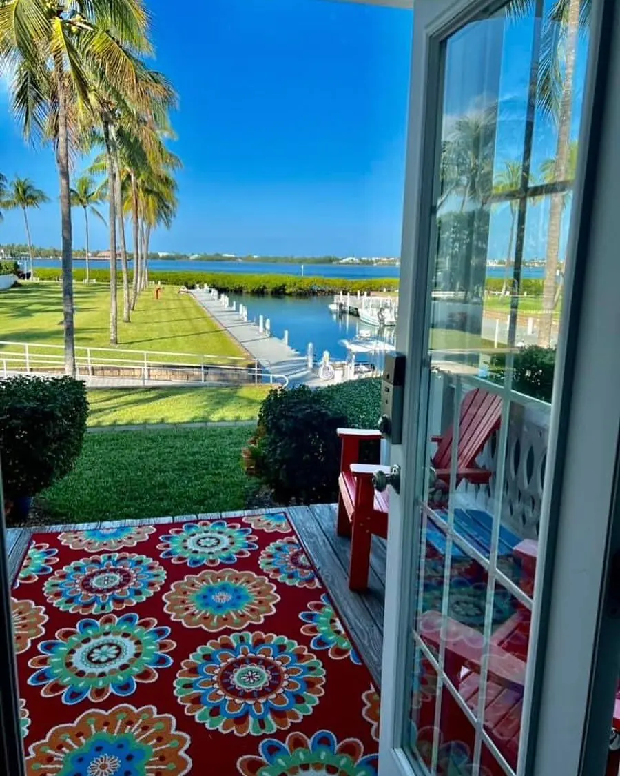 View from inside Indigo Reef #41 through open French doors onto a deck with red Adirondack chairs, a colorful rug, palm trees, and a boat slip on calm water.